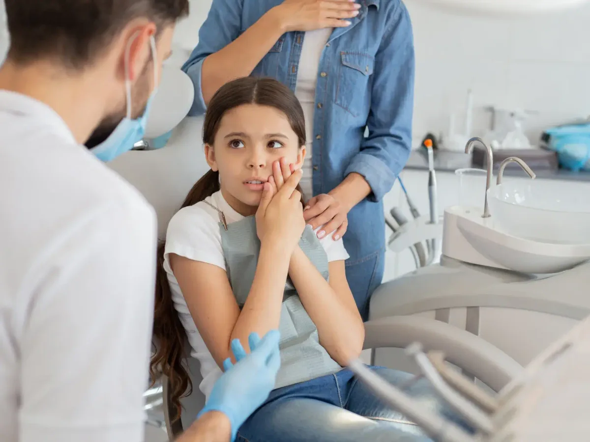 A young patient holding her cheek in discomfort while her mother comforts her at the dental office